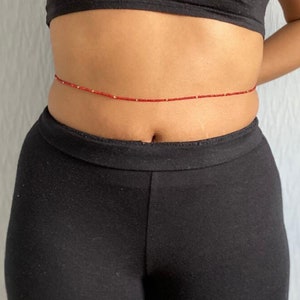 Könnte beinhalten: Ein roter Hüftgürtel mit kleinen goldfarbenen Perlen. Die Hüftperlen werden um die Taille getragen. Die Person auf dem Bild trägt ein schwarzes Crop-Top und eine schwarze Hose.