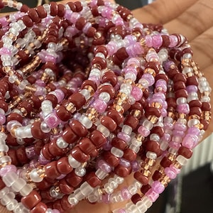 Könnte beinhalten: Nahaufnahme von mehreren Perlensträngen in Burgunderrot, Rosa, Weiß und Gold. Die Perlen sind klein und rund und erzeugen einen strukturierten Effekt. Diese Perlen werden wahrscheinlich für Schmuck oder Dekorationszwecke verwendet.