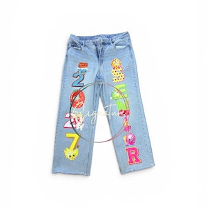 May include: Light blue denim jeans featuring a vertical arrangement of colorful cartoon character graphics and large numbers on the front legs. The left leg displays the number 2027 with various food-themed characters, while the right leg shows the word SENIOR.