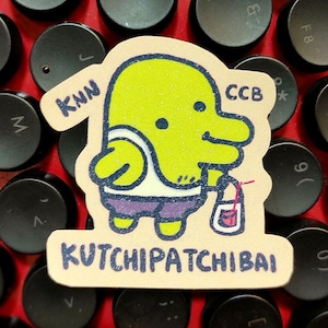 May include: A sticker featuring a cartoon character with a lime green head, wearing a white undershirt and purple shorts, holding a small bag. The sticker has the text "KNN", "CCB", and "KUTCHIPATCHIBAI".