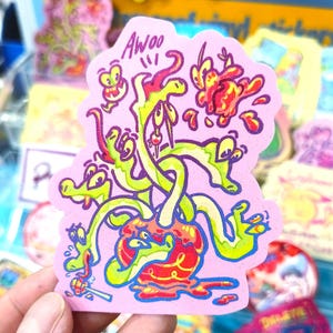 Puede incluir: Una pegatina rosa con un diseño de dibujos animados. La pegatina presenta una mancha central roja y amarilla con apéndices verdes en forma de serpiente y ojos de dibujos animados. La palabra "Awoo" está escrita en la parte superior. La pegatina está sujeta por una mano.