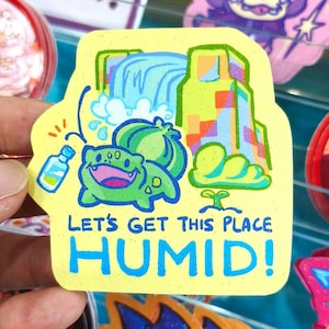 Puede incluir: Una pegatina amarilla con una ilustración de dibujos animados de una criatura verde, una cascada y un edificio. La pegatina tiene el texto "LET'S GET THIS PLACE HUMID!" en azul. También se representa una pequeña botella de agua.