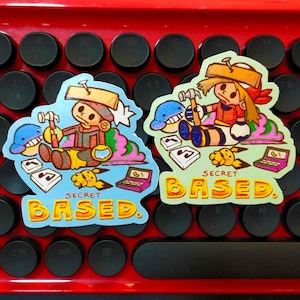 Può includere: Due adesivi colorati con personaggi dei cartoni animati, martelli e la scritta "SECRET BASED". Gli adesivi raffigurano un personaggio con un chiodo in testa, un delfino blu, note musicali e un laptop. Su una tastiera rossa.