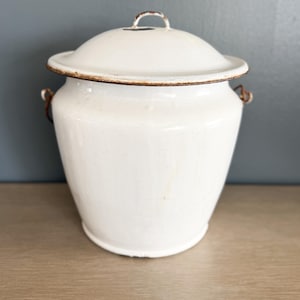 May include: A white enamelware container with a lid. The container has a rounded shape with a lid and two small handles. The rim and handles show signs of rust, adding a vintage aesthetic. The container is sitting on a wooden surface.
