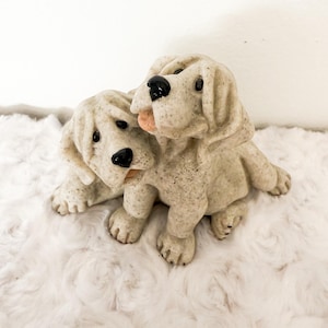May include: A decorative figurine featuring two light beige Labrador Retriever puppies. The puppies are in a playful embrace, with one puppy's tongue sticking out. The figurine has black eyes and noses, and is set on a fluffy white surface.