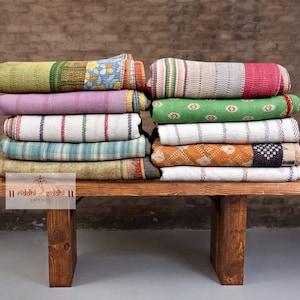 May include: A stack of colorful, rolled-up quilts rests on a wooden bench. The quilts display a variety of patterns, including stripes, floral designs, and geometric shapes. The color palette includes shades of green, pink, white, and orange.