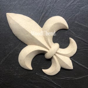 Könnte beinhalten: Ein hellbeiger Holz-Fleur-de-lis-Anhänger mit einer erhabenen, strukturierten Oberfläche. Der Anhänger ist in Form einer stilisierten Lilienblüte mit drei Blütenblättern und einem zentralen Stiel geformt.