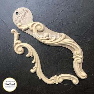 May include: Two unfinished wood decorative scroll ornaments. The ornaments are light beige and feature a curved design with intricate details. The text "WoodUbend" is visible on the attached tag. The bottom left corner has a circular logo with the text "3RD GENERATION WoodUbend IMPROVED DESIGN."