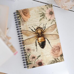 May include: Spiral-bound notebook featuring a large bee illustration with outstretched wings. The bee is centered on a cream-colored background with floral accents and script-like text. The notebook's design includes roses and other flowers in soft pink and peach tones.