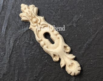 May include: A light-colored, ornate wooden keyhole cover with a decorative floral design. The cover has a keyhole in the center and the text "WoodUbend" is visible. The piece is likely for furniture or decorative use.
