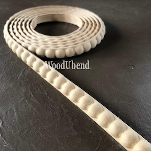 May include: A roll of unfinished wood trim with a repeating ball design. The trim is beige and has the text "WoodUbend" printed on it. The trim is on a dark gray surface.