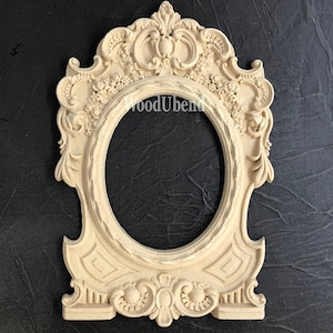 May include: An ornate, off-white wooden frame with a decorative oval opening. The frame features intricate carvings of flowers and leaves, and a central design with a crown.