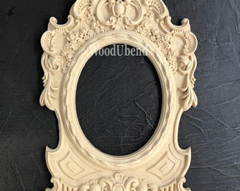 May include: An ornate, off-white wooden frame with a decorative oval opening. The frame features intricate carvings of flowers and leaves, and a central design with a crown.