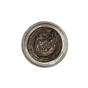 May include: A small, clear glass jar with a brown, creamy substance inside. The jar is empty except for a small amount of the substance left at the bottom.