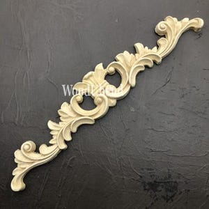 Può includere: Modanatura decorativa in legno color crema, ornata con un design a volute e foglie. La modanatura ha una forma simmetrica ed è posta su uno sfondo scuro e strutturato. Il testo "WoodLand" è visibile sulla modanatura.