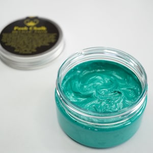 May include: A teal green hair styling cream in a clear glass jar with a black lid. The lid has a gold logo that says "Poet & Skull".