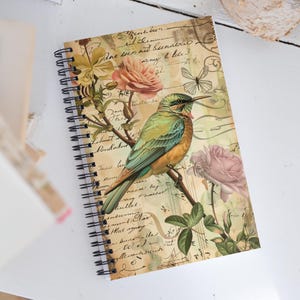 Op de afbeelding: Spiraalgebonden notitieboek met een vintage ontwerp. De omslag toont een vogel op een tak met rozen, tegen een crèmekleurige achtergrond met script- en bloemelementen. De vogel heeft groene, blauwe en oranje veren.