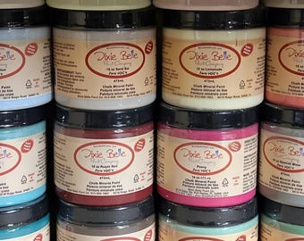May include: A collection of Dixie Belle Chalk Mineral Paint jars in various colors, including Sand Bar, Lemonade, Rustic Red, Peony, and Spanish Moss. Each jar is 16 oz (473ml) and labeled with the brand name and paint color.