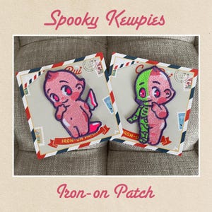May include: Two iron-on patches featuring spooky kewpies. One kewpie is pink with a pink halo and wings. The other kewpie is pink and green with a green halo and wings. Both kewpies have a surprised expression and are holding their hands to their faces. The patches are on a white background with a vintage postage stamp design.