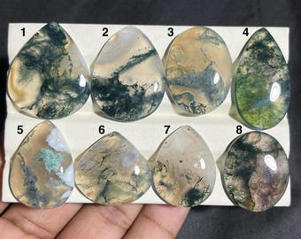May include: Eight polished moss agate gemstones in various shapes, numbered 1 through 8. The stones display a range of colors, including green, white, and brown, with intricate, natural patterns resembling moss or foliage. The gemstones are displayed on a white surface.