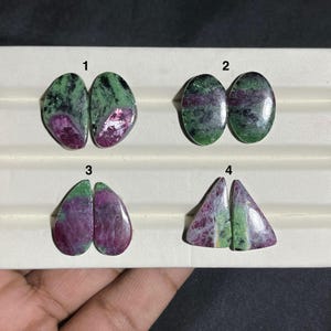 May include: Four pairs of polished ruby in zoisite gemstones. Each pair has a unique shape: oval, teardrop, and triangular. The stones display a mix of green, pink, and purple hues, with visible patterns and textures. The gemstones are displayed on a white surface.