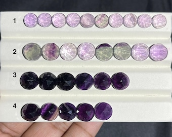 May include: Rows of faceted, round gemstones in shades of purple, lavender, and clear. The gemstones are arranged on a white display. The gemstones vary in color and clarity, with some appearing darker and more opaque than others.
