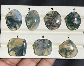 May include: Seven polished moss agate gemstones in various shapes: oval, round, and square. The stones display a mix of green, white, and brown patterns, with a translucent quality. Each stone is unique, showcasing natural inclusions and variations in color.