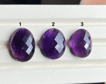 May include: Three oval amethyst gemstones with faceted surfaces are shown. The gemstones are a rich purple hue and have a polished, reflective appearance. They are arranged in a row on a white surface. Measurements are in cm.