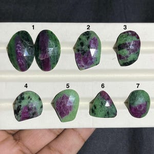 May include: A collection of seven gemstone rings. Each ring features a faceted, oval or teardrop-shaped gemstone with a mix of green and deep red hues. The rings are set on silver bands and displayed on a white surface.