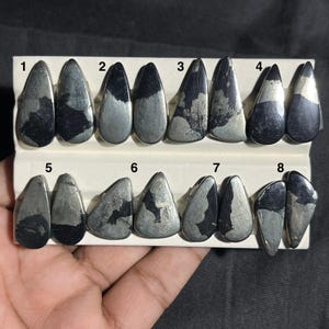 May include: Eight polished pyrite gemstones in dark grey and silver. The gemstones are teardrop and triangular, each with a unique pattern. They are arranged on a white card. Measurements are in centimetres.