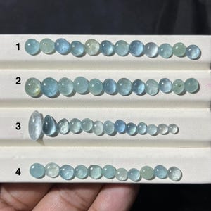 May include: Four rows of aquamarine gemstones in varying sizes and shapes. The gemstones range in color from pale blue to light green. The stones are oval, round, and teardrop-shaped, displayed on a white surface. The gemstones are arranged in a gradient of sizes.