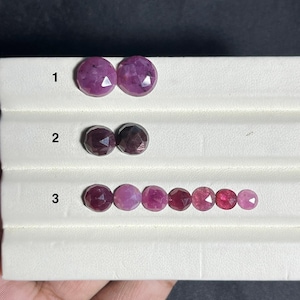 Puede incluir: Una colección de piedras preciosas redondas y facetadas de color rosa y rojo oscuro dispuestas en filas sobre un expositor blanco. La fila 1 tiene dos piedras rosas, la fila 2 dos piedras rojo oscuro y la fila 3 siete piedras en tonos variados.