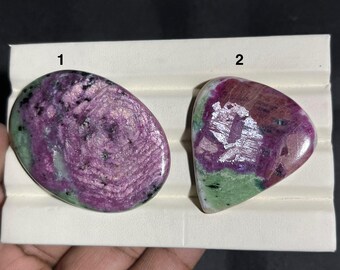 Può includere: Sono esposte due pietre preziose lucide, una ovale e una triangolare. La pietra ovale è principalmente viola scuro con inclusioni verdi e nere. La pietra triangolare ha una tavolozza di colori simile, con un mix di viola, verde e argento.