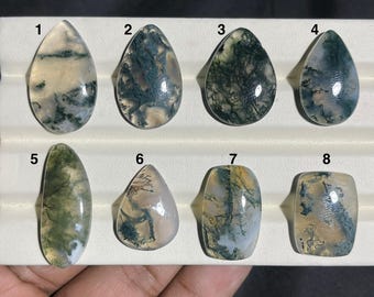 May include: Eight moss agate gemstone cabochons in various shapes, including teardrop and oval. The stones display green, white, and brown patterns. Each stone is set on a ring base, showcasing the natural beauty of the gemstones.