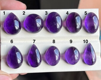 May include: Ten polished amethyst gemstones in various shapes, including teardrop and oval. The gemstones are a deep purple colour and are displayed on a white surface. The gemstones are numbered 1 through 10.