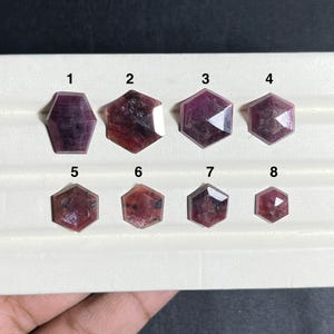 May include: Eight faceted, hexagonal-cut, deep pink and reddish-purple gemstones arranged in two rows on a white display tray. Each stone is numbered from one to eight, showcasing varying sizes and internal inclusions within the crystal structure.