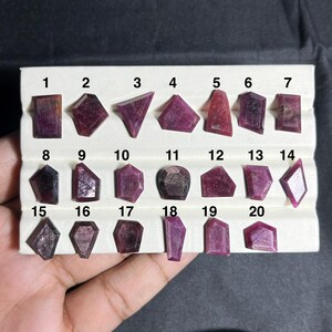 Puede incluir: Una colección de veinte piedras preciosas de rubí facetadas, dispuestas en tres filas sobre una bandeja de exhibición blanca. Cada piedra está numerada del 1 al 20, mostrando varias formas geométricas irregulares y tonos rojo oscuro.