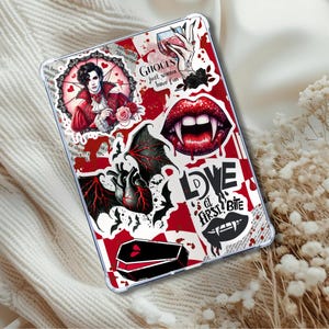 Puede incluir: Una funda transparente para tableta con un diseño de vampiro en rojo y blanco. La funda presenta ilustraciones de un vampiro, labios con colmillos, un corazón con alas de murciélago y el texto "GHOULS just wanna have fun" y "LOVE at first bite".