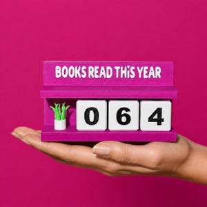 May include: A magenta book counter with the text "BOOKS READ THIS YEAR" displayed on top. The counter shows the number "064" on white blocks. A small potted plant is on the left side of the counter. The counter is held in a hand against a magenta background.