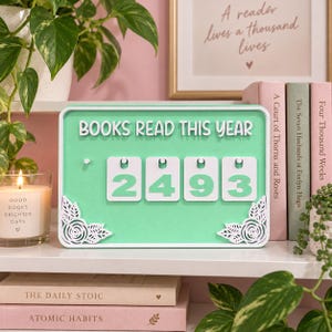 May include: A rectangular mint green sign with white borders and floral corner accents. The text "BOOKS READ THIS YEAR" is printed at the top. Four white square tiles with mint green numbers "2", "4", "9", and "3" are displayed in the center.