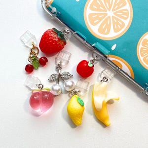 Puede incluir: Un conjunto de charms de frutas coloridas, que incluyen una fresa, una cereza, una manzana, un melocotón, un limón y un plátano. Los charms están unidos a soportes de carcasa de teléfono de plástico transparente.