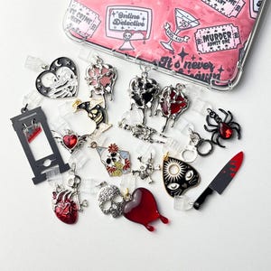 Puede incluir: Una colección de colgantes de temática gótica en varias formas, incluyendo corazones, calaveras y una araña. Los colgantes son negros, rojos y plateados, con una funda transparente para teléfono en el fondo con el texto "Online Detective".