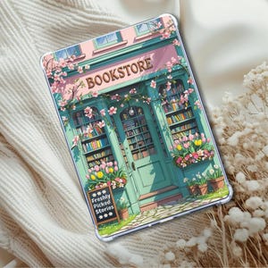 May include: A clear tablet case featuring a whimsical illustration of a bookstore. The artwork depicts a teal storefront with the word "BOOKSTORE" above the entrance, surrounded by pink blossoms and flower boxes. A sign reads "Freshly Picked Stories."