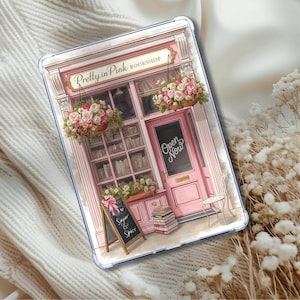 Peut inclure: Une coque de tablette transparente avec une illustration rose d'une librairie. La librairie, nommée "Pretty in Pink Bookshop", a une porte rose avec le texte "Open Now", des paniers de fleurs et une enseigne "Sugar & Spice".