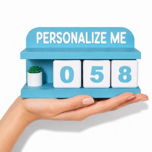 May include: A blue decorative block with the text "PERSONALIZE ME" and the numbers "058". A small potted plant sits on the left side. The block is held in a hand.