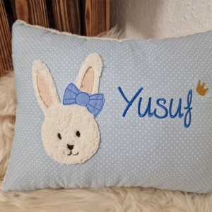 Könnte beinhalten: Ein hellblaues Kissen mit weißen Punkten zeigt ein gesticktes Häschen mit einer blauen Schleife und den Namen "Yusuf" mit einer goldenen Krone.