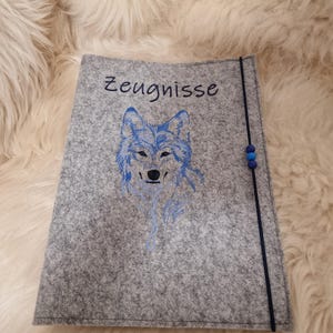 Könnte beinhalten: Ein grauer Filz-Dokumentenhalter mit dem Wort "Zeugnisse" in schwarzer Schrift. Ein blaues gesticktes Wolfsdesign ist mittig auf der Vorderseite. Ein schwarzes Gummiband mit blauen Perlen sichert den Halter.