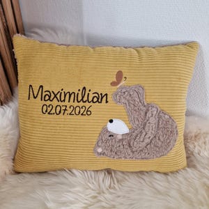 Könnte beinhalten: Ein rechteckiges Kissen aus senfgelbem Kord mit einem gestickten braunen Teddybären, der liegt, und einem kleinen Schmetterling darüber. Der Text "Maximilian" und "02.07.2026" ist in schwarzem Garn auf der linken Seite des Kissens eingestickt.