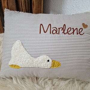Könnte beinhalten: Ein rechteckiges Kissen mit beigefarbenem und weißem horizontalem Streifenmuster. Das Kissen ist mit dem Namen Marlene in brauner Schreibschrift bestickt, gefolgt von einem kleinen braunen Herz. Eine weiße, strukturierte Gänseapplikation mit gelbem gepunktetem Schnabel.