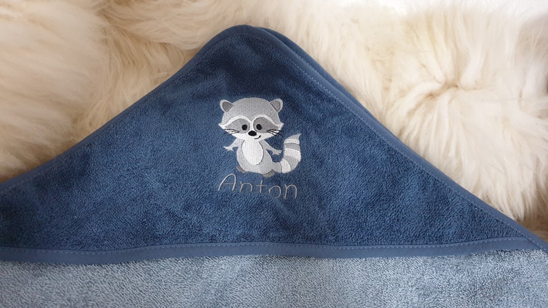 以下が含まれることがあります： アントンという名前が刺繍された、アライグマの刺繍が施された青いフード付きタオルです。